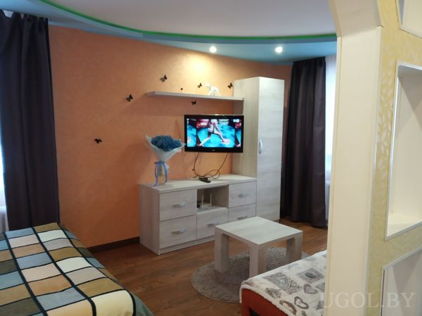 Современная гостиная с телевизором на светлой тумбе, вазой с синими цветами, оранжевой стеной с бабочками и диваном сбоку.