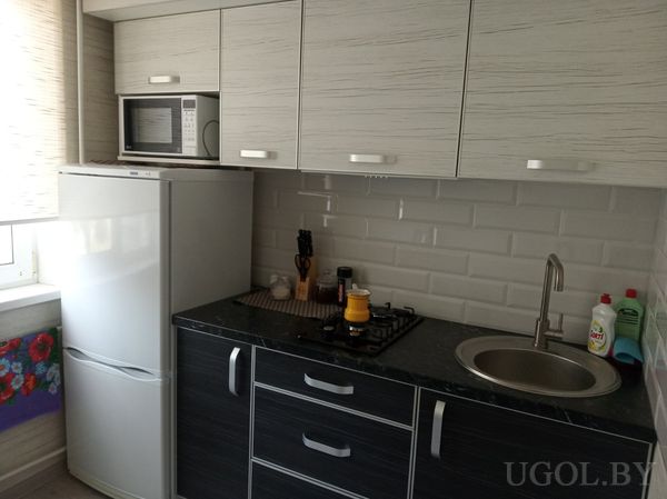 Компактная кухня с плитой, жёлтой кастрюлей, микроволновкой на холодильнике, мойкой и белым фартуком из плитки.