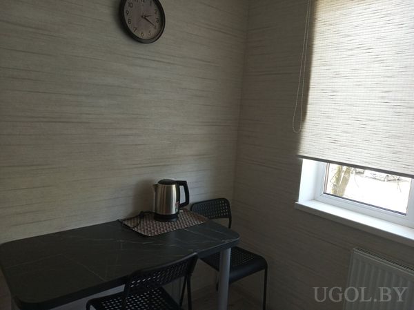 Небольшая столовая с чёрным столом, электрическим чайником, двумя пластиковыми стульями и окном с бежевой шторой.