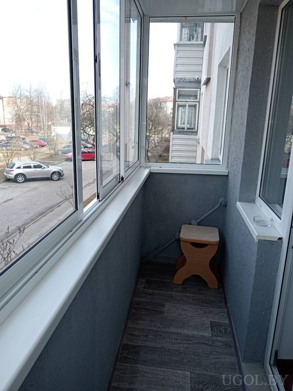 Узкий застеклённый балкон с тёмным полом, серыми стенами, деревянным табуретом и видом на двор с машинами.