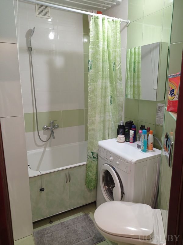 Компактная ванная с зелёной плиткой, ванной с занавеской, стиральной машиной, унитазом и зеркальным шкафчиком.