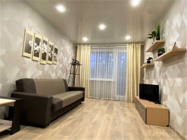 Светлая гостиная с бежевым диваном, тремя картинами с цветами на стене и большим окном с занавесками.