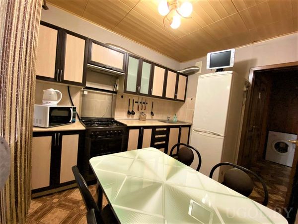 Кухня с деревянными шкафами, плитой, холодильником и обеденной зоной с круглым стеклянным столом и чёрными стульями.