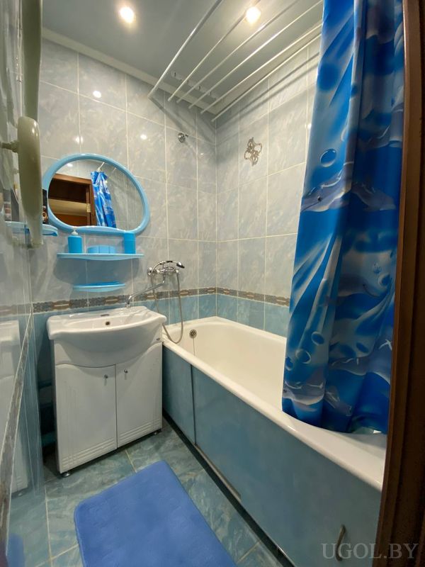 Компактная ванная с белой ванной, голубой плиткой, круглым зеркалом и синим ковриком.