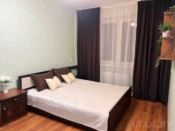 Двуспальная кровать с симметрично разложенными подушками, светло-зелёные стены с золотыми звёздами, тёмные шторы и полка с растениями.