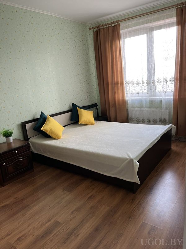 Современная спальня с кроватью в тёмной раме, белым покрывалом, зелёными и жёлтыми подушками, зелёными стенами и окном с тюлем.