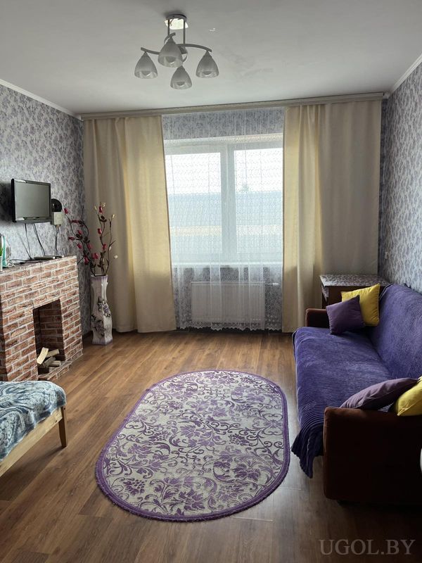 Уютная гостиная с фиолетовым диваном, жёлтыми подушками, декоративным камином, телевизором и ковром с цветочным узором.