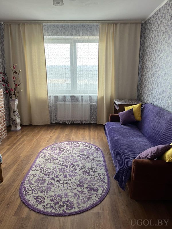 Тёплая гостиная с диваном в фиолетовом покрывале, подушками, вазой с ветками и большим окном с тюлем и шторами.