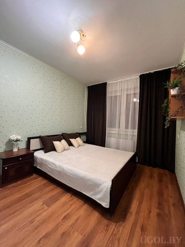 Белое покрывало, декоративные подушки, тумбочка с цветами, ламинированный пол, два настенных стеллажа с растениями и тёмные шторы.