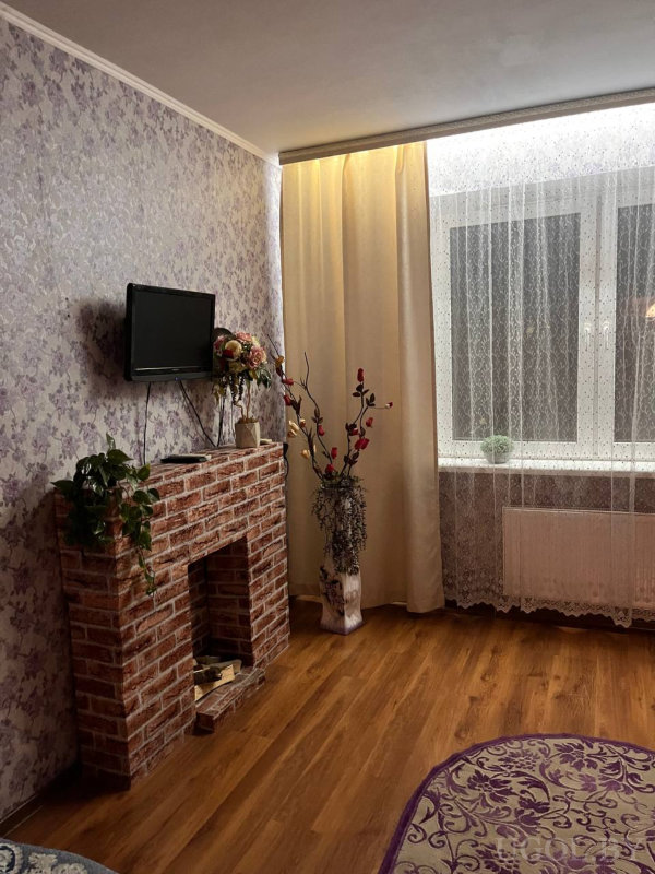 Камин с растениями и ТВ на фоне фиолетового цветочного полотна, окно с кремовыми шторами и высокая декоративная ваза с ветками.