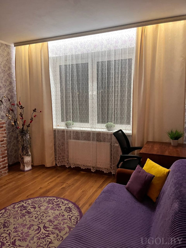 Большое окно с тюлем и бежевыми шторами, диван с подушками, круглый ковёр, деревянный пол, рабочий стол с чёрным креслом и растениями.