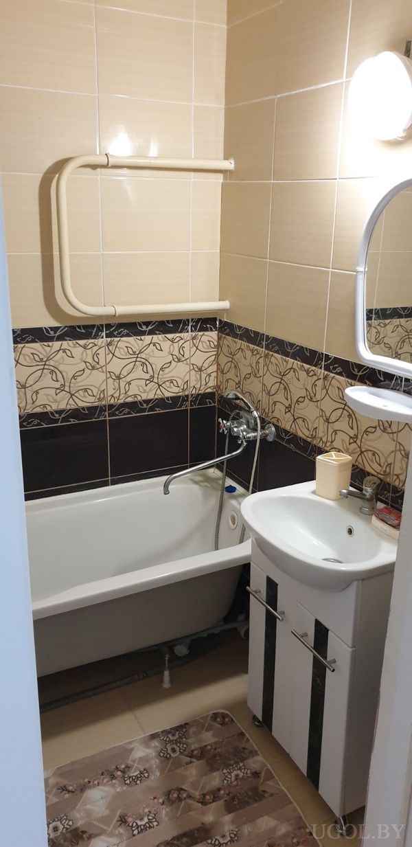 Ванная с белой ванной, тёмной плиткой с узором, зеркалом над раковиной и светильником, рядом — коврик и шкафчик.