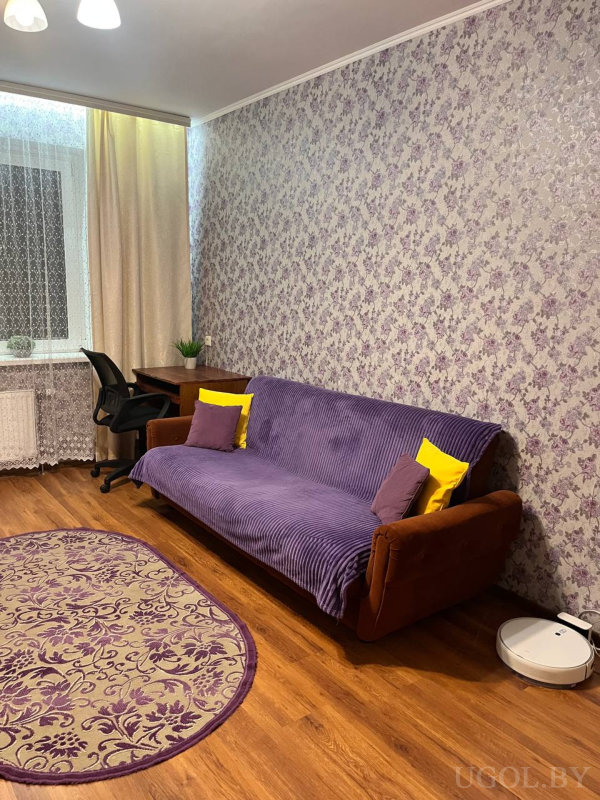 Диван с фиолетовым покрывалом и жёлто-фиолетовыми подушками, рабочая зона у окна, фиолетово-бежевый ковёр и робот-пылесос на полу.