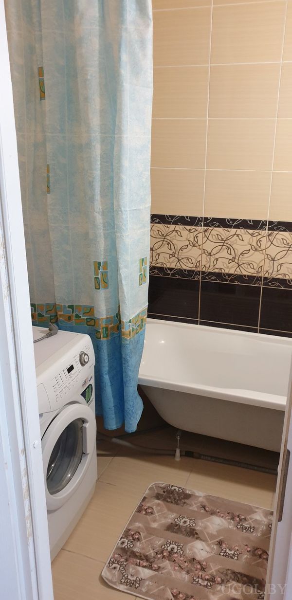 Компактная ванная с белой ванной, синей занавеской с узором, стиральной машиной и ковриком с цветочным рисунком.