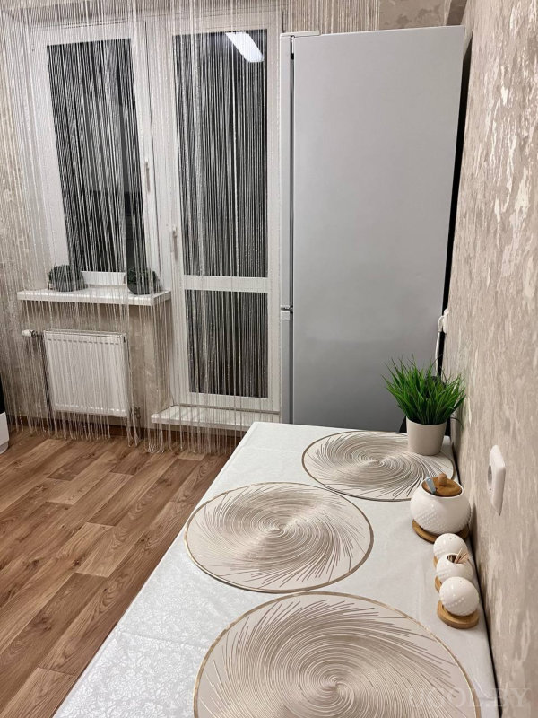 Белый стол со скатертью, спиральные салфетки, горшок с зеленью, набор керамики, серебристый холодильник и длинные нитяные шторы у окна.