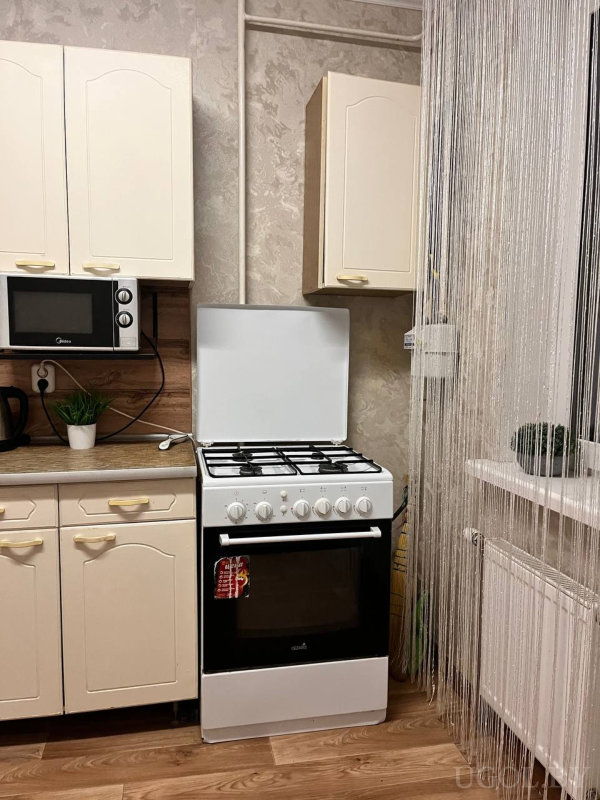 Белая газовая плита с полотенцем, микроволновка «Midea», кремовые шкафы, чайник, растение и нитяная штора, за которой радиатор и подоконник.