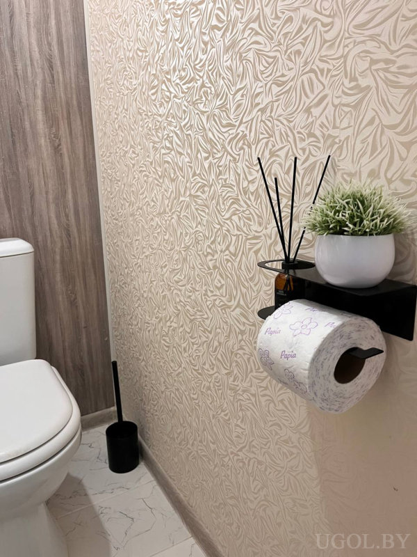 Белый унитаз, чёрный ёршик, деревянная панель, светлая фактурная стена с листовым узором, полка с «Papia», растением и диффузором.