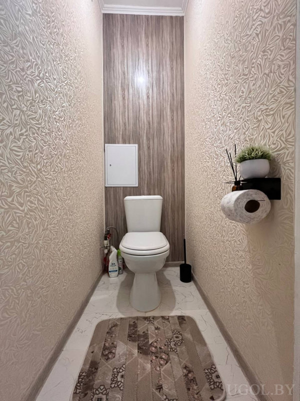 Центрально расположенный унитаз, светлые фактурные стены, чёрная полка с бумагой и диффузором, декоративный коврик и чистящие средства у основания.