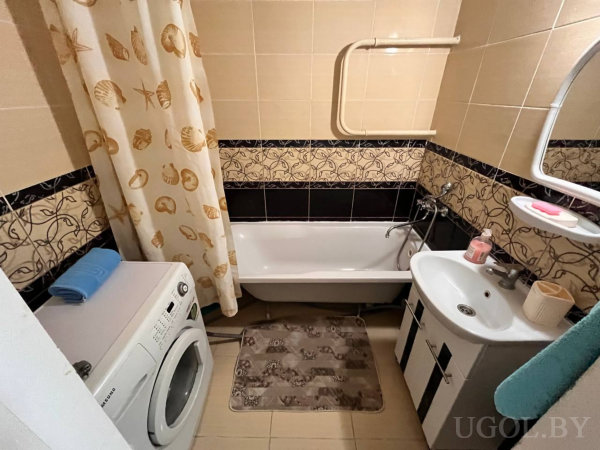 Белая ванна с шторой в морской тематике, тумба с раковиной и зеркалом с полкой, розовая мыльница и стиральная машина с голубым полотенцем.