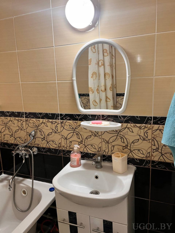 Овальное зеркало с полкой, тумба с раковиной, розовый дозатор, цветочный бордюр по плитке, вид на ванну с душем и синее полотенце.