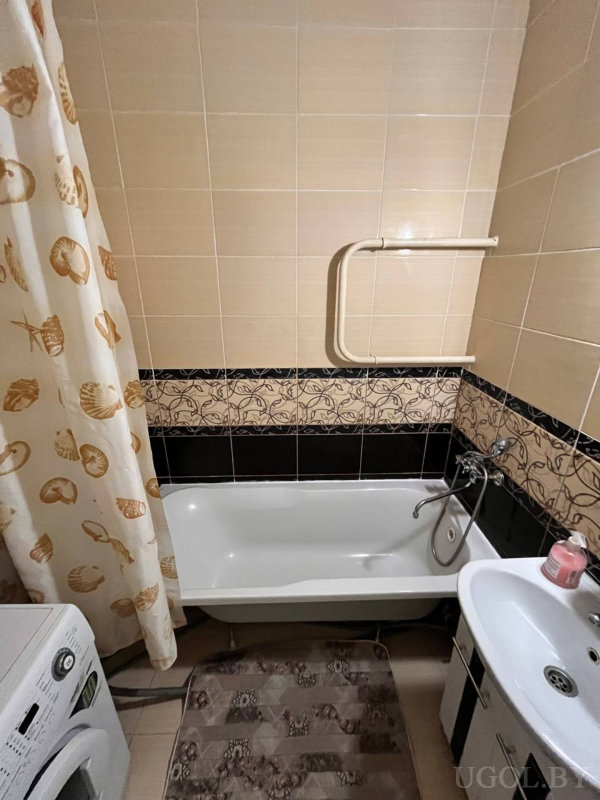 Белая ванна с настенным поручнем, шторка с ракушками, тумба с раковиной и розовым дозатором, коричневый коврик и стиральная машина сбоку.