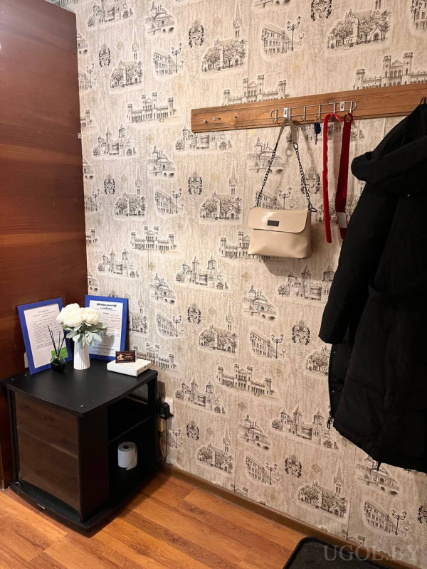 Настенная вешалка с сумкой, пальто и лентой на фоне обоев с европейскими зданиями. Ниже — чёрная тумба с полками, на ней ваза с цветами, карточки и документы в рамках.