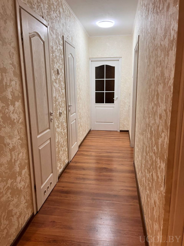 Деревянный пол, бежевые текстурные обои, потолочный светильник и белые двери, включая одну с остеклением. Пространство выглядит чисто и упорядоченно.