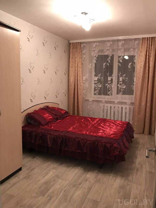 Уютная спальня с двуспальной кроватью, красным сатиновым покрывалом, светлым гардеробом и окнами с цветочным тюлем.