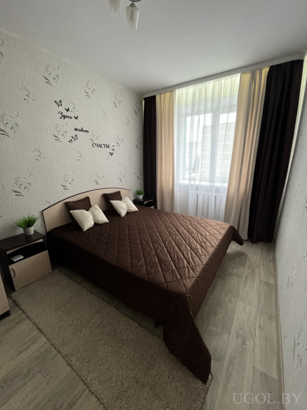 Современная спальня с двуспальной кроватью, коричневым покрывалом, растениями на тумбочках и декоративной надписью на стене.