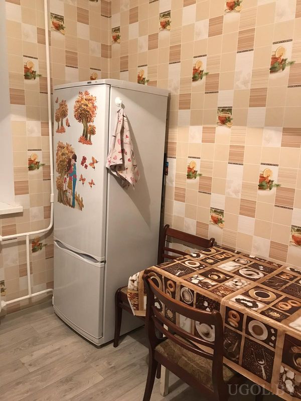 Уютный кухонный уголок с белым холодильником, украшенным наклейками, столом с кофейной скатертью и тремя деревянными стульями.