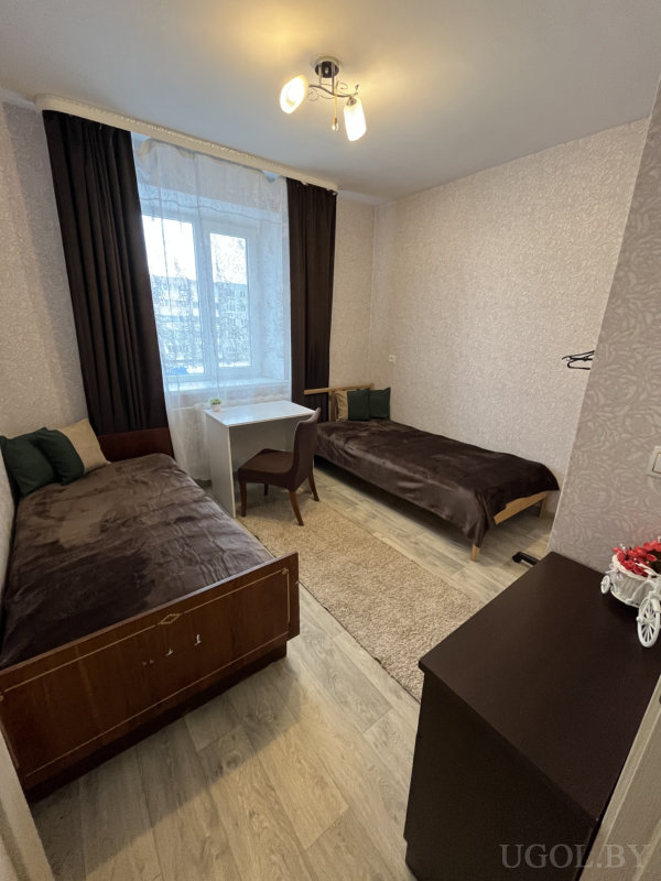 Интерьер с двумя кроватями, белым столом и декоративной тележкой с цветами.