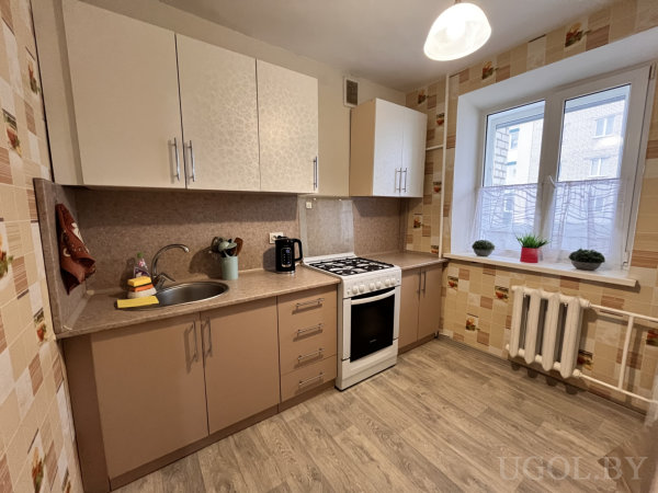 Современная кухня с белыми и бежевыми фасадами, растениями на подоконнике и плиткой с рисунками.