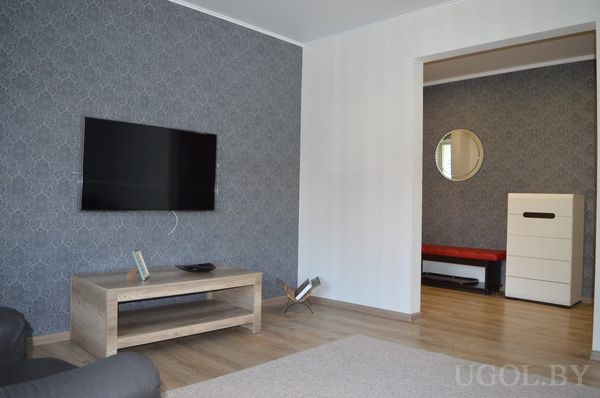 Минималистичная гостиная с телевизором на тёмной стене, светлым кофейным столиком и видом в соседнюю комнату с зеркалом и красной банкеткой.