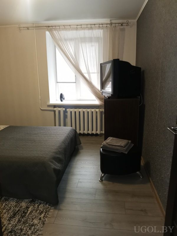 Небольшая спальня с кроватью в сером покрывале, окном с белыми шторами, радиатором и телевизором на деревянной тумбе.