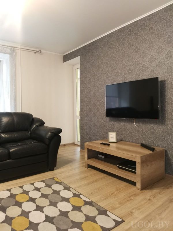 Современная гостиная с чёрным креслом, телевизором на тёмной стене, светлой тумбой и ковром с геометрическим узором.