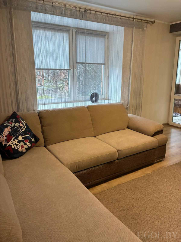 Бежевый диван с цветной подушкой, нитяной тюль и ролеты на окне с видом на деревья.