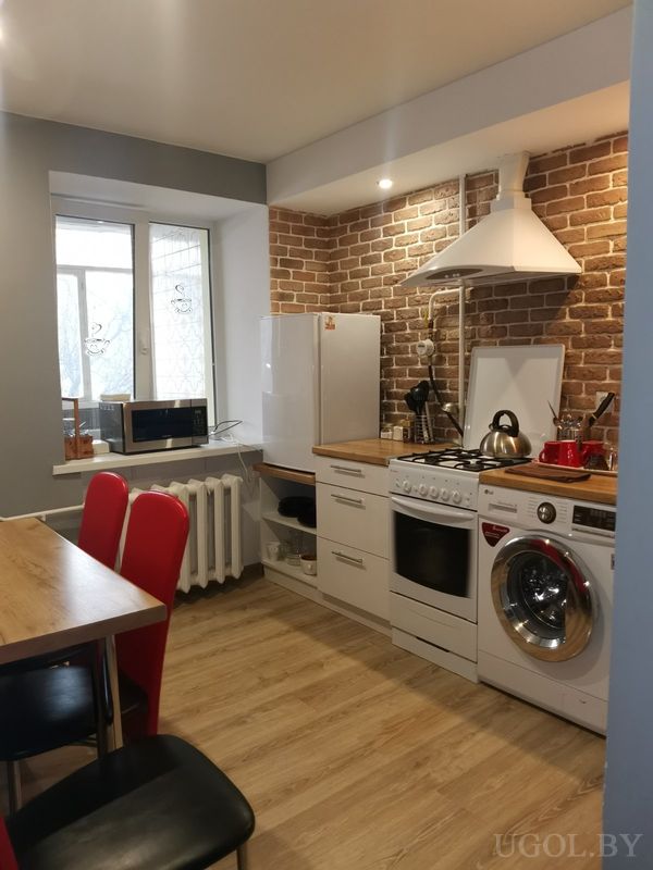 Компактная кухня с белым холодильником, плитой, стиральной машиной, микроволновкой у окна и кирпичным фартуком.