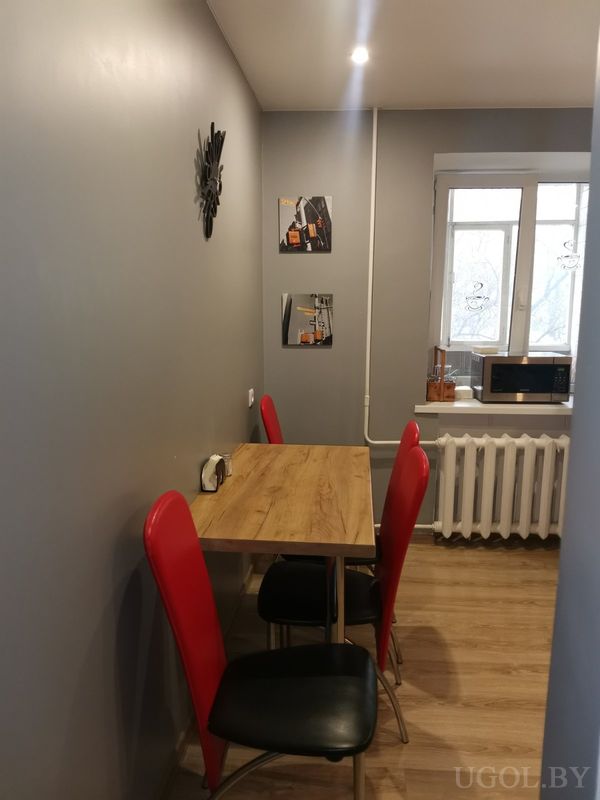 Небольшая столовая с деревянным столом, четырьмя стульями с красными спинками, серой стеной и часами над картиной.