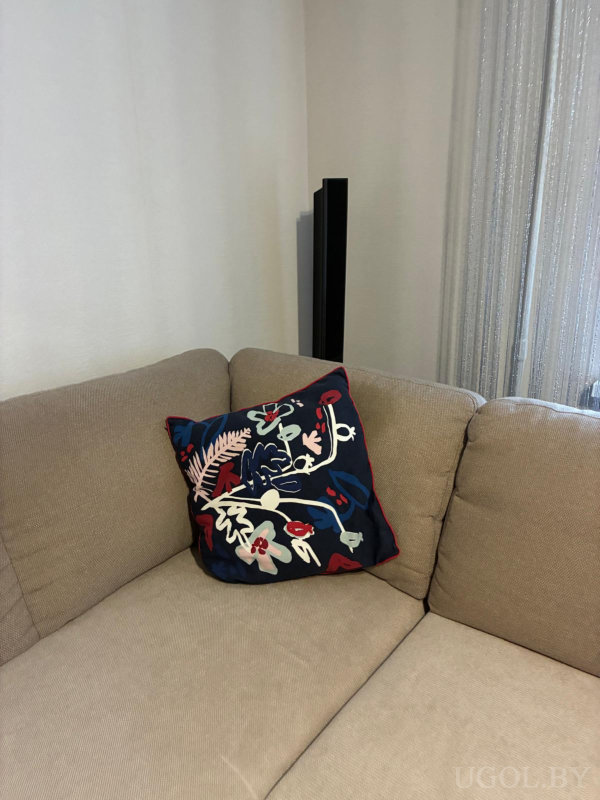 Бежевый диван в углу комнаты, подушка с абстрактным цветочным рисунком и колонка у стены.