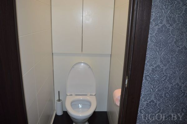 Компактный санузел с белым унитазом, шкафчиком над ним, розовой бумагой и белой плиткой.