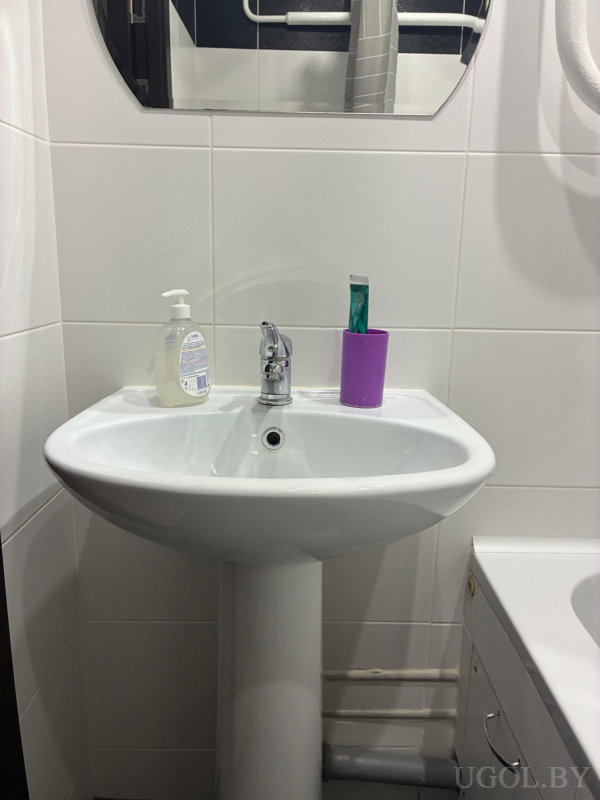 Белая раковина с мылом и зубной щёткой, зеркало с изогнутым краем и плитка на стене.