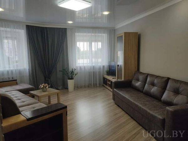 Современная гостиная с тёмно‑коричневым кожаным диваном, журнальным столиком с цветами, телевизором на тумбе и большими окнами с белыми шторами.