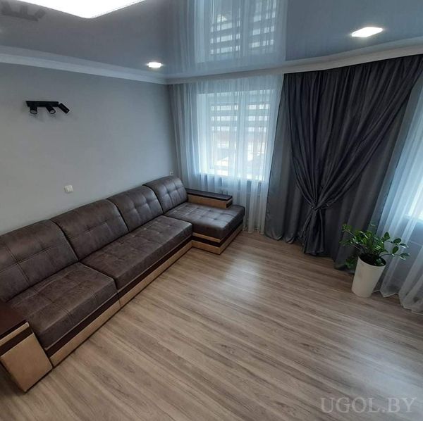 Минималистичная гостиная с угловым коричневым диваном, пуфом, большим окном с белыми шторами и серыми стенами.