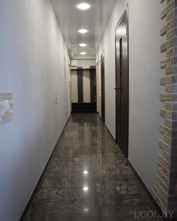 Современный коридор с тёмным глянцевым полом, белыми стенами, встроенной подсветкой и шкафом с зеркалом в конце.