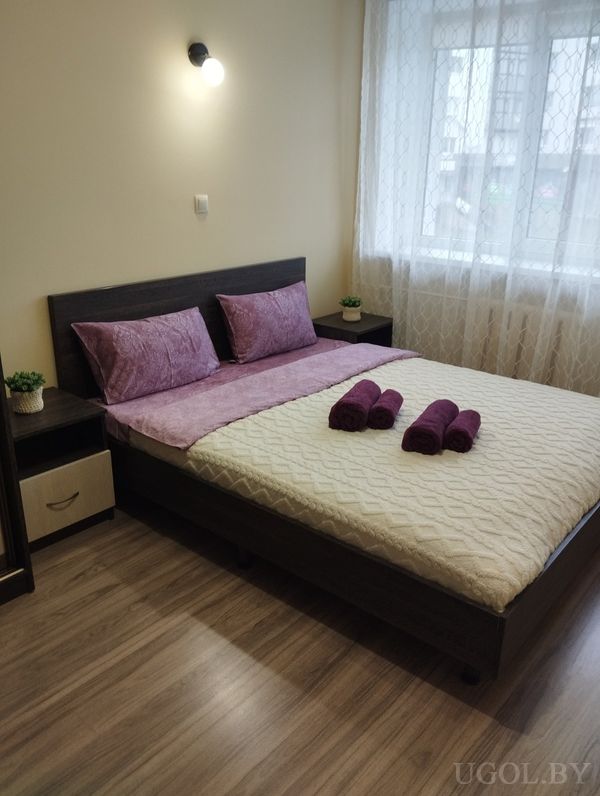 Уютная спальня с двуспальной кроватью, кремовым покрывалом, фиолетовыми подушками и полотенцами, прикроватными тумбами и растениями.