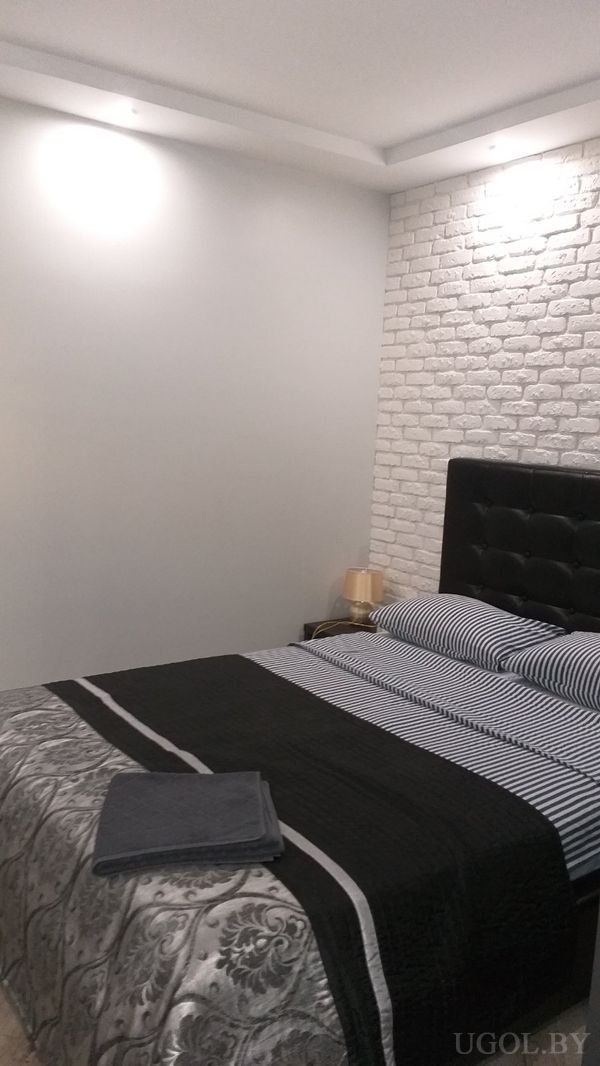 Минималистичная спальня с кроватью в чёрном изголовье, чёрным покрывалом с узором, белой кирпичной стеной и прикроватной лампой.