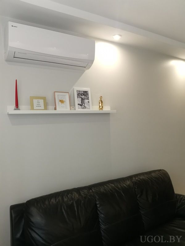 Светлая гостиная с чёрным диваном, белой полкой с декором, кондиционером над ней и серой стеной.