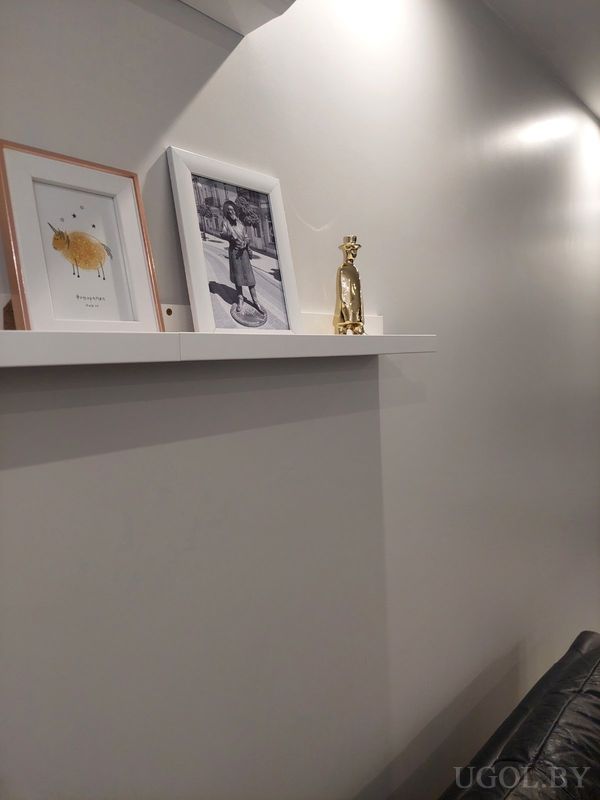 Светлая стена с белой полкой, на которой стоят рамки с фото и фигурка жирафа, рядом чёрный диван.