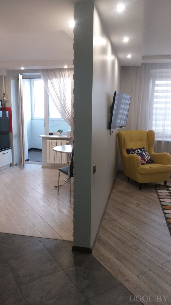 Современные апартаменты с жёлтым креслом, телевизором на стене, ковром с узором и обеденной зоной за перегородкой.