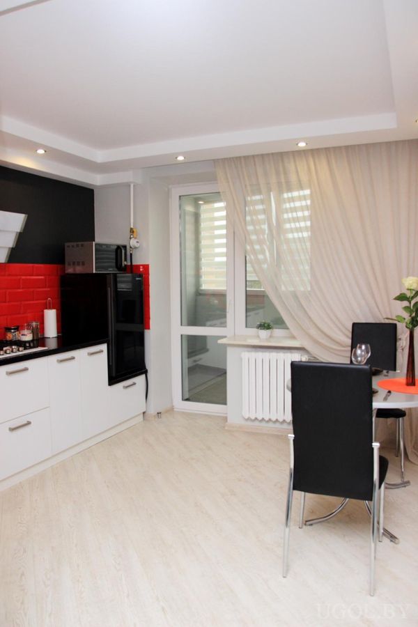 Современная кухня с белыми шкафами, чёрной столешницей, красным фартуком и встроенной техникой.